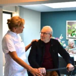MoverFlexi. Man zit op Mover Flexi, verzorger staat naast hem met haar hand op zijn schouder