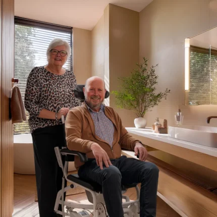 Anytoilet M douche-toiletstoel in een badkamer thuis, een zorgvrager zit in de stoel en zijn mantelzorger staat erachter