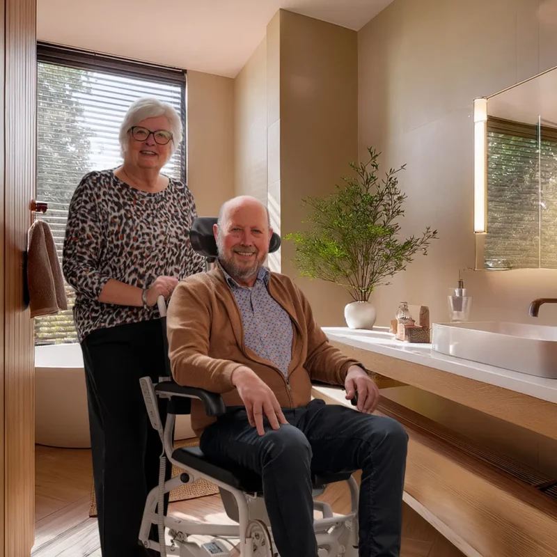 Anytoilet M douche-toiletstoel in een badkamer thuis, een zorgvrager zit in de stoel en zijn mantelzorger staat erachter