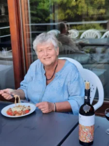 Mayke met kort wit haar zit buiten aan een donkerblauw tafeltje en eet pasta met tomaten. Voor haar staat een wijnfles met kleurrijk etiket. Op de achtergrond een terras met witte stoelen en groen.