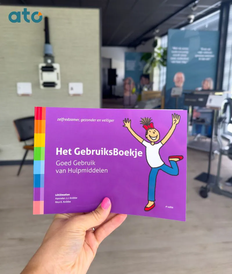 Hand houdt Het GebruiksBoekje van LOCOmotion vast, met op de paarse cover de titel Goed Gebruik van Hulpmiddelen, in het ATO Experience Centre.