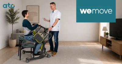 eMove Family. Een jonge man komt met een sta-oplift uit een stoel omhoog in zijn woonkamer, een zorgverlener bedient het hulpmiddel en staat naast hem.