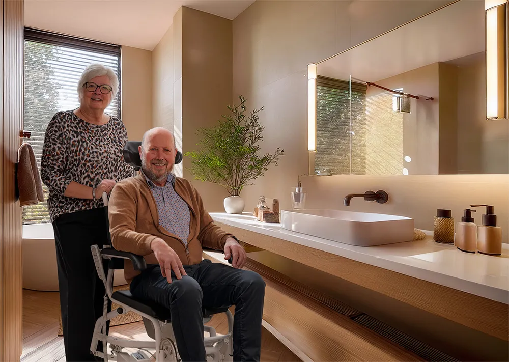 Anytoilet douche-toiletstoel in een badkamer, een man zit in de stoel bij de wastafel en een vrouw staat achter hem