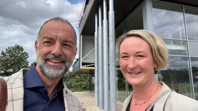 Selfie van Wibo en Sabine, lachend buiten voor het modern gebouw van ATO met een glazen gevel en stalen kolommen
