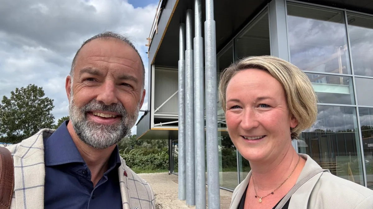 Selfie van Wibo en Sabine, lachend buiten voor het modern gebouw van ATO met een glazen gevel en stalen kolommen