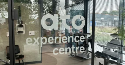 De glazen toegangsdeur van het ATO Experience Centre in Gorredijk, met het logo ato experience centre op het glas. Achter het glas staan hulpmiddelen opgesteld in de showroom, met daglicht uit de ramen aan de achterzijde.