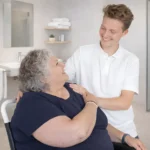 Solid douche-toiletstoel in een badkamer, een zorgvrager met overgewicht zit in de stoel en een zorgverlener staat achter haar